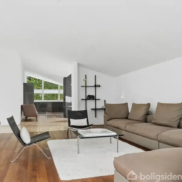 En stue med en stor beige sofa og to moderne stole på et lyst tæppe. Stuen har hvide vægge og trægulv, og i baggrunden ses et rum med spisebord.