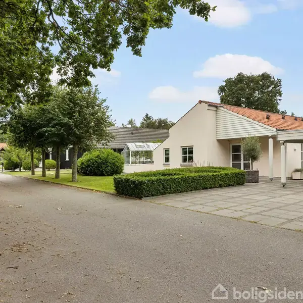 En hvid bungalow står stille ved en rolig vej omgivet af grønne træer og buske. Huset har et fladt tag og en indkørsel med flisebelægning.