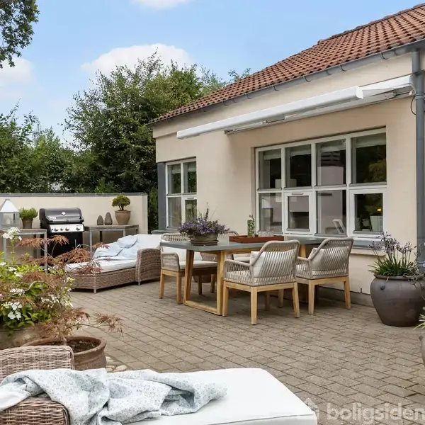 Terrasse med træbord og stole arrangeret til spisning, omgivet af blomsterkrukker og en grill i hjørnet. En mur og grønne træer danner baggrunden. Ingen tekst.