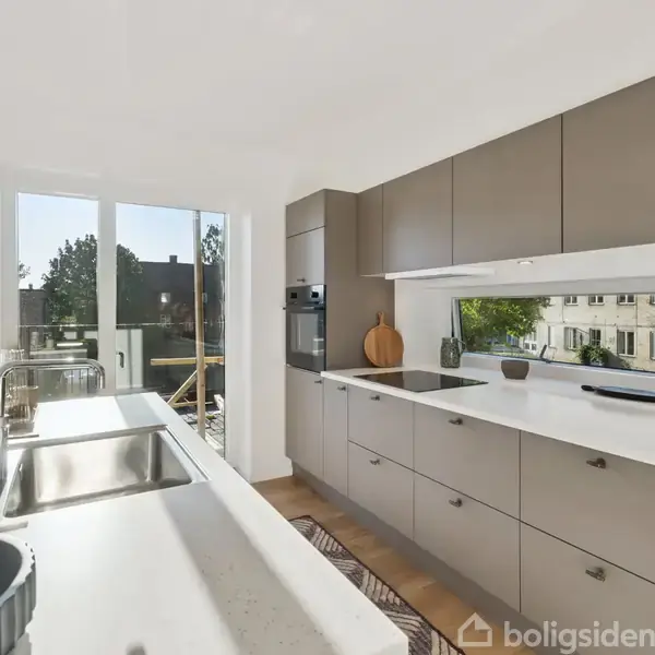 Køkken med moderne, grå skabe og hvid bordplade, har vask og ovn indbygget. Vinduet giver udsigt til et solrigt udendørsmiljø med træer og bygninger i baggrunden.
