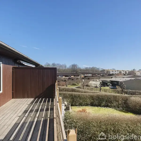 Terrasse med træpaneler strækker sig fra hus under blå himmel, omgivet af grønne hække og udsigt til beboelsesområde med huse i baggrunden.