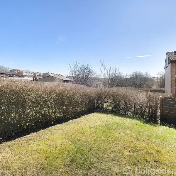 Græsplæne ligger stille omgivet af tætte hække, mens en murstensbygning står til højre under en klar blå himmel. Axelgade i baggrunden er delvist synlig gennem træerne.