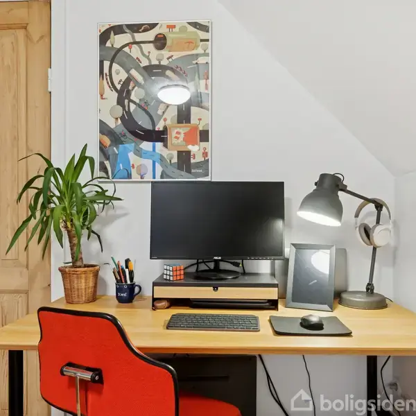 Computerskærm står på et skrivebord ved siden af en lampe og en plante. Baggrunden består af en trækdør og en abstrakt kunstplakat på væggen.