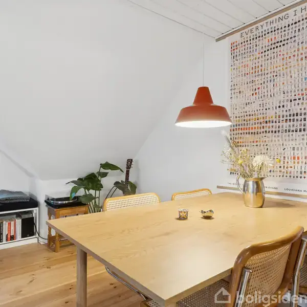 Et træbord med stole omkring står i en lys spisestue. En plakat med teksten "EVERYTHING I HAVE" hænger på væggen. Der er en plante og en guitar i hjørnet.