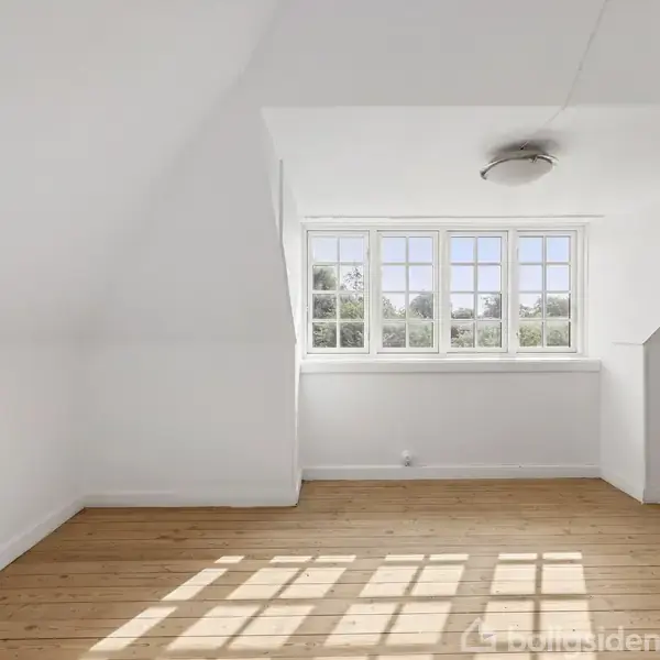 Et lyst rum med trægulv, hvor solen skinner igennem et stort vindue, skaber skygger på gulvet. Væggene og loftet er hvide; en rund lampe hænger fra loftet.
