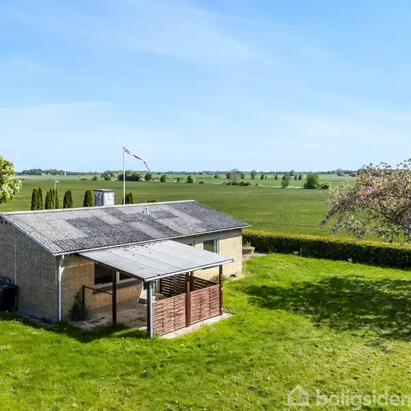 Et enkelt, lille hus med fladt tag står på en græsplæne. Et træ og en flagstang ses ved siden af, omgivet af åbne marker under en klar blå himmel.