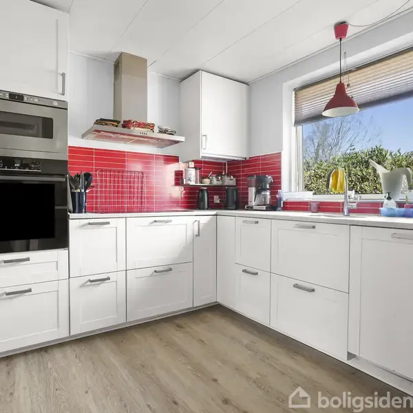 Hvidt køkken med ovn og mikrobølgeovn indbygget i skabene, lys trælaminatgulv, røde fliser som backsplash, køkkenredskaber organiseret på bordpladen, samt et vindue med udsigt til grønne buske.