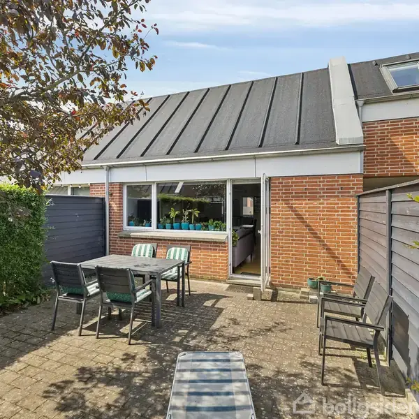 En terrasse med havemøbler står foran et murstenshus med sort tag. Døren til huset er åben, og der er buske og hegn omkring terrassen.