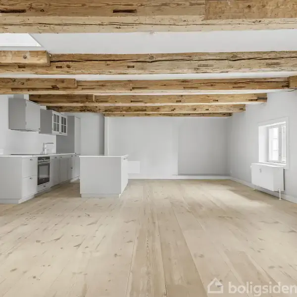 Køkken-alrum med moderne hvide køkkenelementer til venstre. Trægulve strækker sig over rummet, mens synlige træbjælker i loftet tilføjer karakter. Store vinduer lyser den rummelige, minimalistiske indretning op.