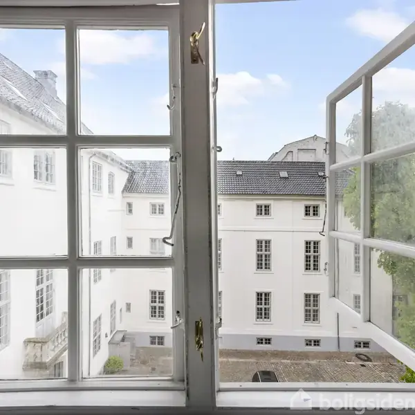 Et åbent vindue viser en udsigt til en indre gård omgivet af hvidmalede bygninger med mange vinduer. Himlen er klar og blå.