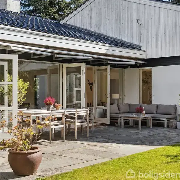 Terrassemøbler stående udenfor en hus, omfatter et bord og stole ved åben terrasse med grønne planter og potter omkring. Sollys skinner på græsset, der omgiver stenbelægningen.