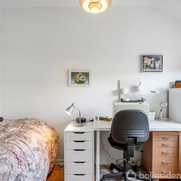 Skrivebord med stol og lampe står i et lyst værelse ved siden af en seng med blomstrede tæpper. Væggene har små malerier, og reolen er fyldt med små genstande.