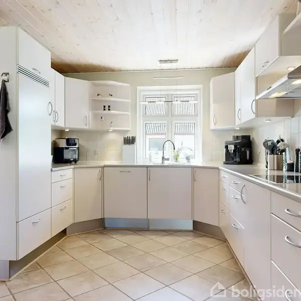 Et lyst køkken har hvide skabe og skuffer i et L-formet layout, med en vask under et vindue. Køkkenredskaber og små apparater står på bordpladen.