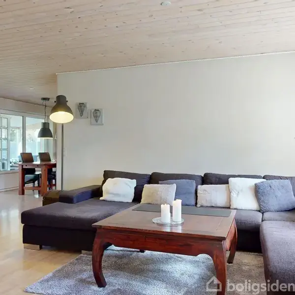 En stue indeholder en brændeovn, flankeret af en reol med brænde. Et ur hænger på væggen over en kommode. Et billede og levende lys står på en skænk.