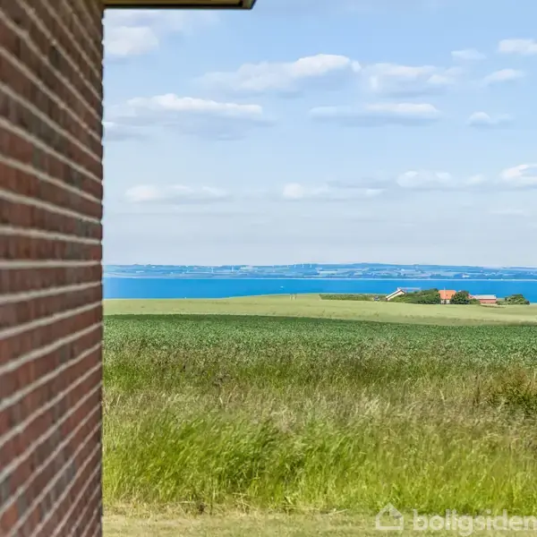 Et murstenshus med solpaneler på taget ligger i en grøn have med en udsigt over havet i baggrunden. Klar blå himmel med få skyer.