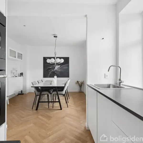 Et køkken-alrum med spisebord og seks stole. Køkkenbordet har vask og indbygget ovn. Store vinduer slipper lys ind, mens et maleri og lampe hænger over spisebordet.