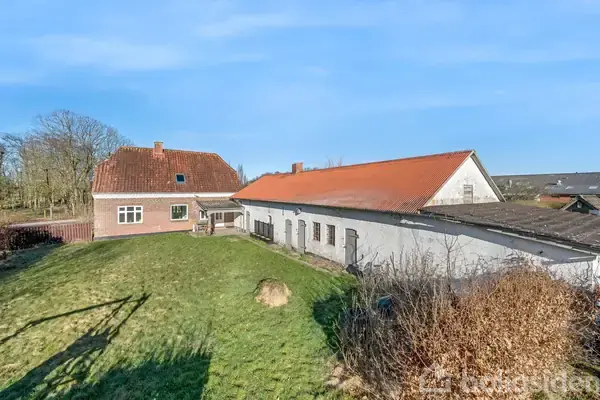 Hus med rødt tegltag og sidelænge i landligt område, omgivet af græs og buske, under en klar blå himmel.