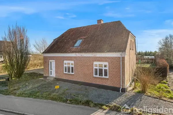 Rødt murstenshus står på en grøn grund med skrånende tag og kvistvindue. En hvid dør og hvide vinduesrammer vender mod vejen i forstadsmiljø.