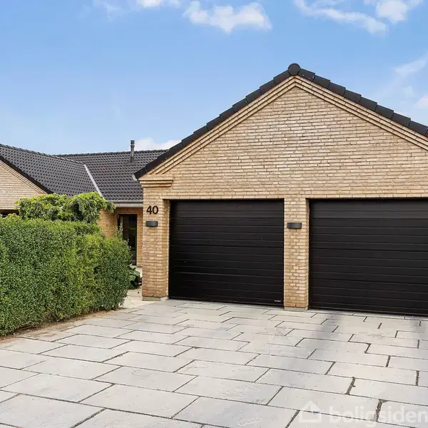 Et murstenshus med to sorte garageporte, placeret ved siden af en flisebelagt indkørsel. Husrække nummer 40 vises tydeligt. Omkring huset er der grønne hække og en klar blå himmel.