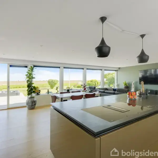 Et moderne køkken-alrum med en stor ø i midten, to sorte pendel lamper hænger over. Store vinduer viser en udsigt til en terrasse og grønne omgivelser udenfor.