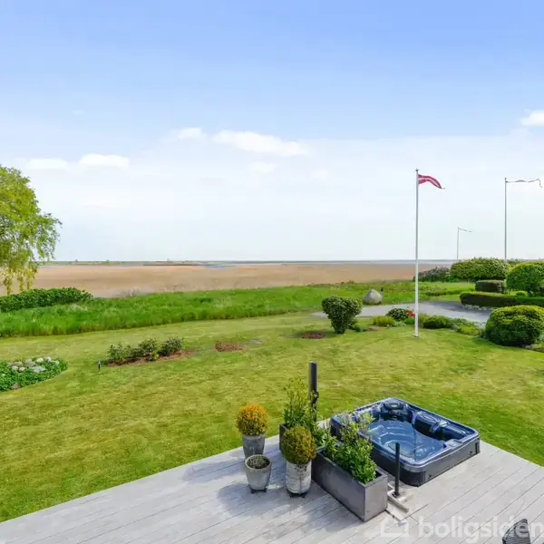 Boblebadet står på en træterrasse og er omgivet af planter, mens en flagstang med Dannebrog er rejst i en velplejet have med udsigt over en mark og himmel.