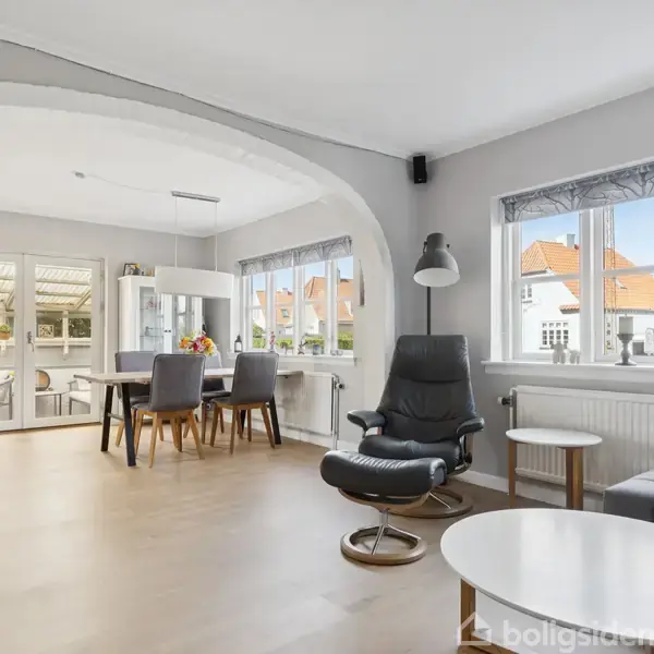 Stuen har et lyst trægulv og er indrettet med moderne møbler, herunder en lænestol, sofabord og spisebord. Store vinduer giver udsigt til nabohuse og masser af naturligt lys.