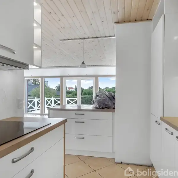 Et moderne køkken med hvide skabe står stille. Baggrunden viser store vinduer med udsigt til en grøn, udendørs have. Loftet er i lyst træ, som matcher det minimalistiske design.