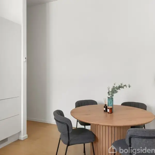 Rundt bord med plante og glasvase står i et moderne spiseområde med fire sorte stole, ved siden af hvidt skab og lys væg. En plante i hjørnet og billede på væggen.
