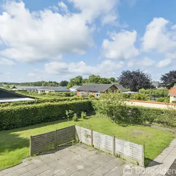 Rødt murstenshus med sort tag står i en velholdt have. Fliser danner en indkørsel og terrasse foran huset. Træhegn omkranser ejendommen, og himlen er blå med skyer.