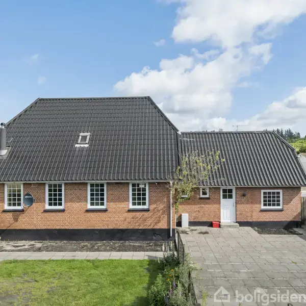 En baghave med en græsplæne og et flisebelagt område er omkranset af lave hække. Området er omgivet af lave huse med blå himmel og spredte skyer i baggrunden.
