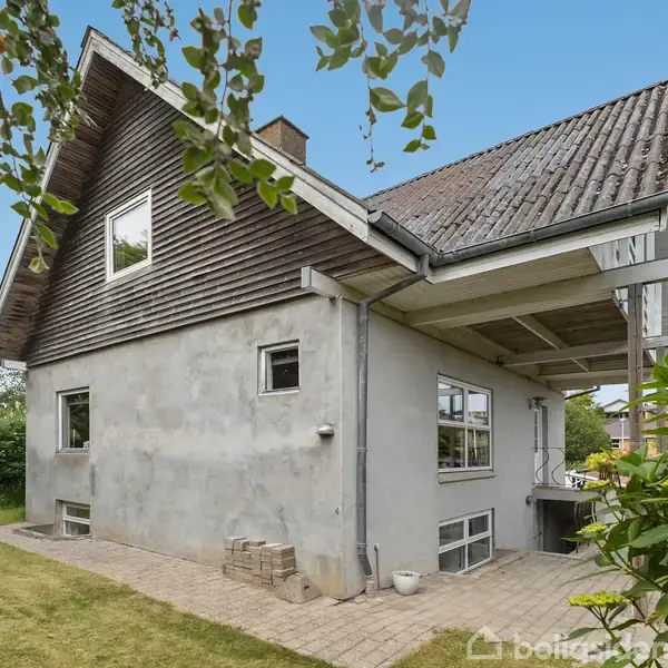 Et to-etagers hus står stille blandt grønne omgivelser, med en pudset facade og træbeklædning. En stenbelagt sti omkranser huset, og et træet tagoverhæng giver skygge.