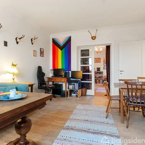 Et værelse med en stor sofa og sofabord til venstre, et skrivebord med computer-station foran en regnbuevæg i midten, og et spisebord med stole til højre. Væggene har kunst og hjorteopsatser.