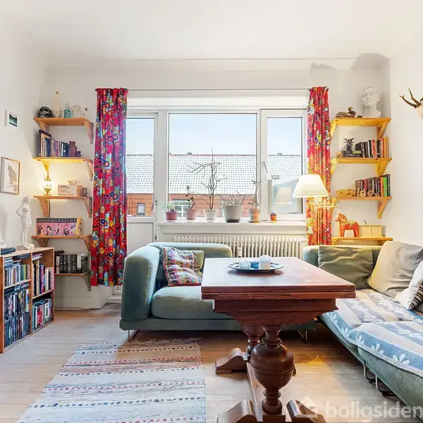 En hyggelig stue med en blå sofa og et træbord står midt i rummet. Vindue med farverige gardiner, reoler med bøger og lamper i baggrunden. Tæpper ligger på gulvet.