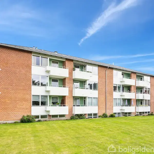 En tre-etagers murstensbygning med hvide altaner står stille på en grøn plæne, omgivet af flere træer under en klar blå himmel.