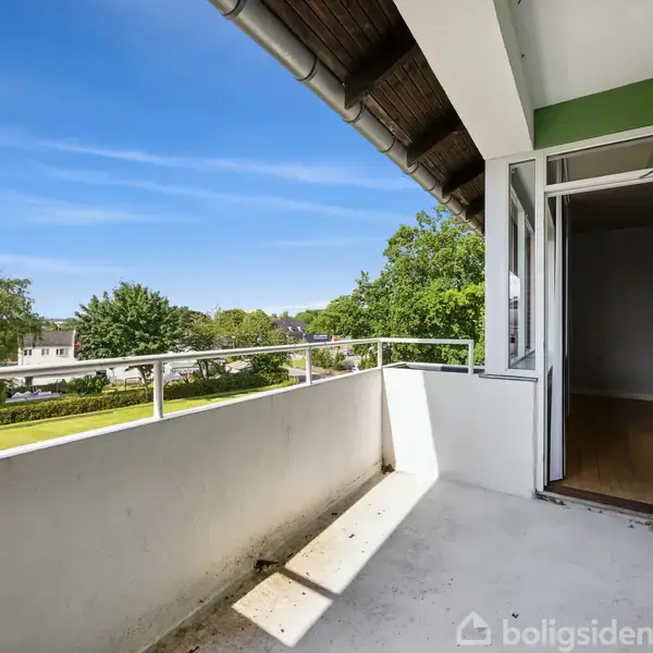 Altan med hvidt gelænder åbner mod en blå himmel. En dør står åben til en indendørs stue. Udsigt til grønne træer og huse i det fjerne.