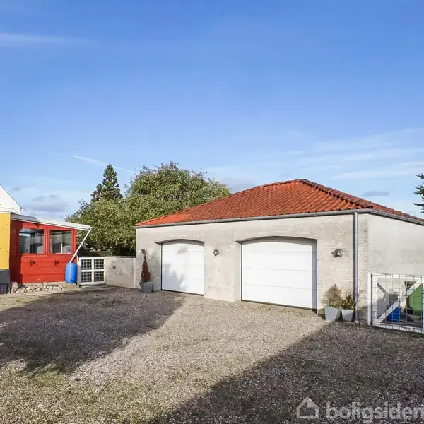 Grå garage med to hvide porte står på en grusbelagt gårdsplads. Ved siden af står et gult hus med en rød tilbygning med vinduer. Omkring er der træer og blå himmel.