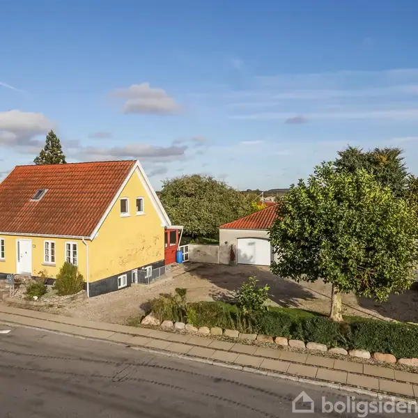 Gul murstenshus med rødt tegltag, omkranset af en have med grussti. Til højre ses en garage og et stort træ. Baggrunden inkluderer flere huse under en blå himmel.