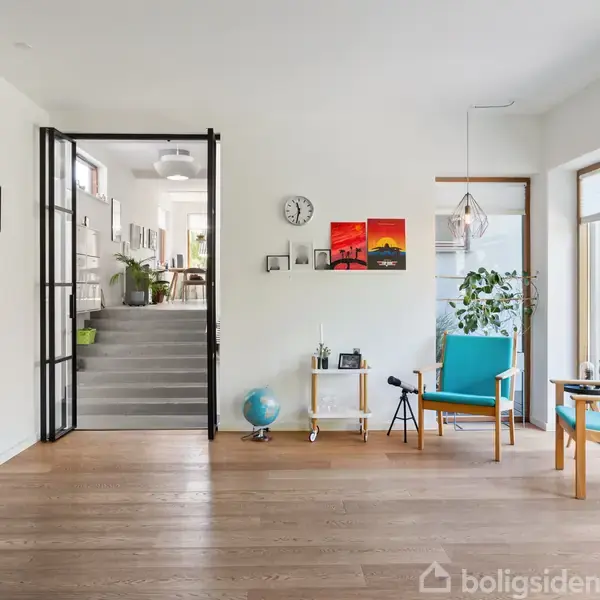 Stue med træplankegulv og moderne møbler, herunder to blå stole og en væghængt reol. En sort glasdør fører til et køkken. Væggene har kunst og ure.