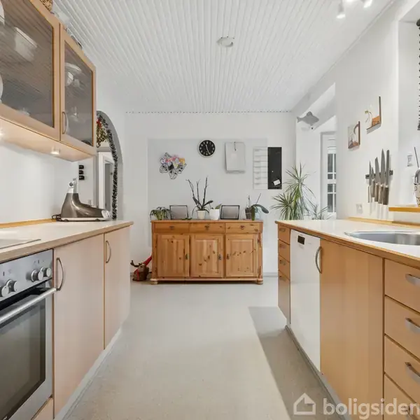 Et moderne køkken med lyse træskabe og stålkomfur, omgivet af hvide vægge. På bordet står en knivholder ved siden af en vask. En kommode findes i baggrunden.