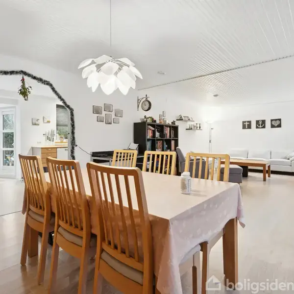 Spisebord med otte træstole står i et lyst rum. En moderne lysekrone hænger over bordet. I baggrunden ses en stue med sofa, reol og et dekoreret døråbning.