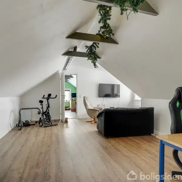 Et loftsrum med trælaminatgulv indeholder en motionscykel, sofa, vægmonteret tv og skrivebord. Grønt pynt hænger fra loftsbjælker. Et åbent dørhul fører til et andet rum.