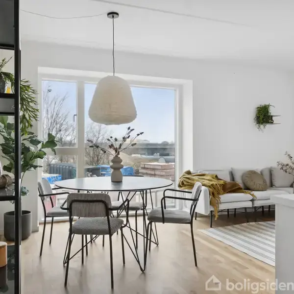 Spisebord med stole står i et lyst rum med trægulv og stor vinduesparti, der fører ud til en balkon. En sofa med tæppe og planter dekorere hjørnet.