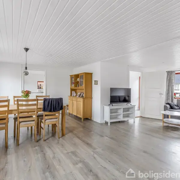 Spisestuemøbler med træbord og seks stole står i et rummeligt, lyst rum med trægulv. En reol står mod væggen; et tv er placeret på et hvidt møbel.