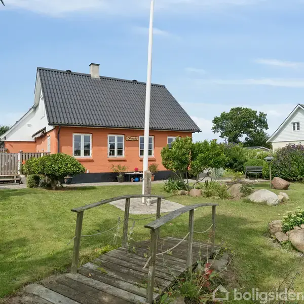Et orange hus står stille i en velholdt have med en lille træbro i forgrunden. I baggrunden ses en anden bolig omringet af buske og træer under en klar himmel.