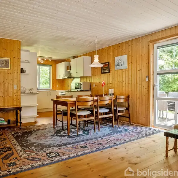 Spisebord med stole står på et tæppe i en træbeklædt stue. Rummet har åbent køkken med hvide skabe, en dør til en terrasse og grøn udsigt. Væglamper og billeder er på væggen.