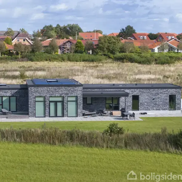 En moderne, gråstensvilla står statisk med solpaneler på taget i et grønt område. Bagved ses rødt tagede huse og træer i en landsby.