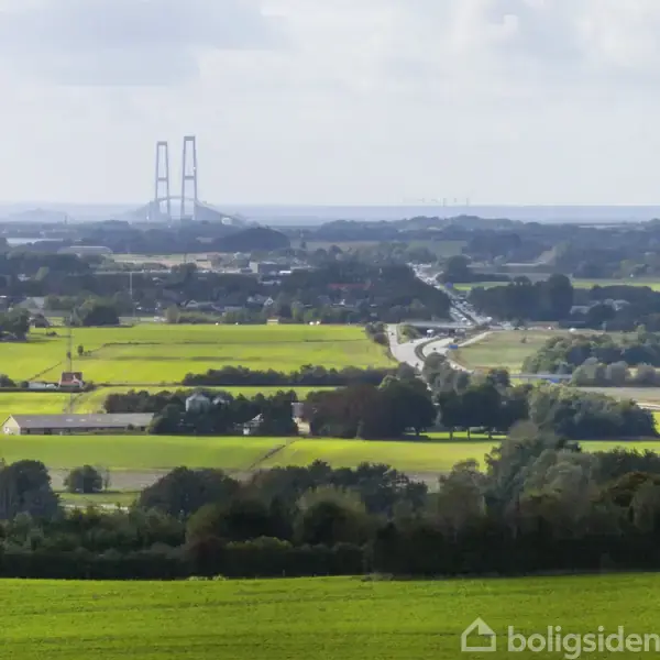 Bro i horisonten strækker sig over grønne marker og en snoet vej, med skove og åbne landskaber i baggrunden.