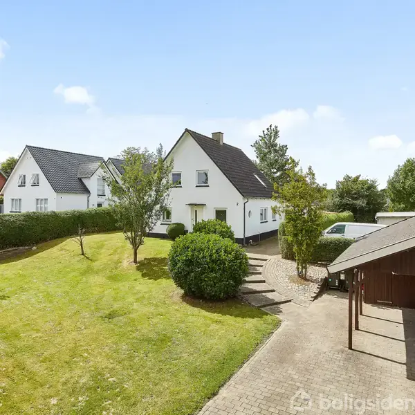 Hvide huse med sorte tage står i en velplejet have med en flisebelagt indkørsel. En carport med trætag er til højre, omgivet af buske og træer.