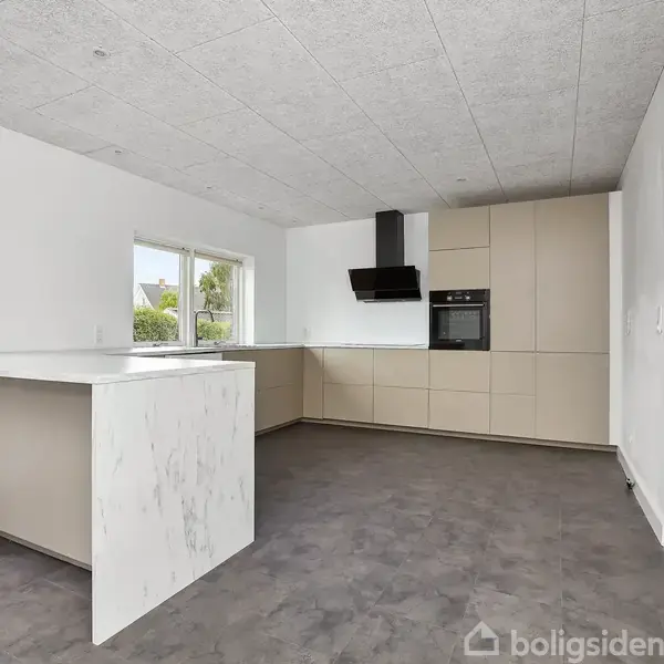 Et moderne køkken med hvide vægge og grå gulvfliser. Køkkenet har et marmorinspireret bord og beige skabe med indbygget ovn. To store vinduer giver udsigt til en have.