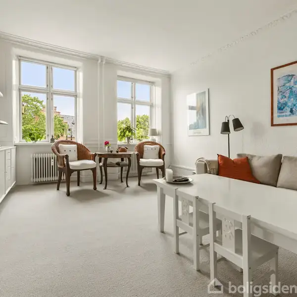 Stuen har to stole og et lille bord foran tre store vinduer med udsigt til grønne træer. Sofa med puder og lampe i højre side. Maleri på væggen.